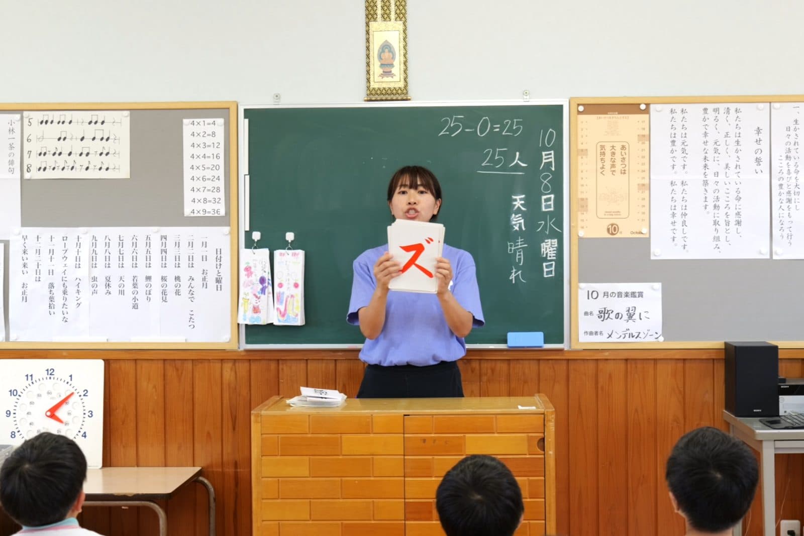 日課活動で、先生がカタカナのフラッシュカードを掲げ、子どもたちが声を合わせて読む様子。黒板には今日の日付・天気と計算が書かれている