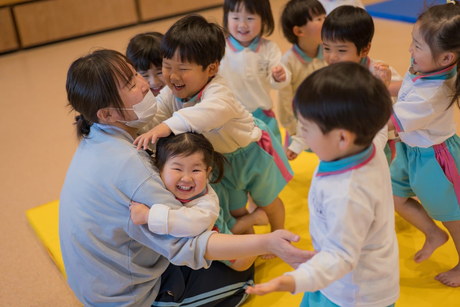 マットの上で、笑顔の子どもたちに抱きつかれる先生